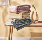 pile de trois plaid pliés, un rose sur un jaune sur un gris, sur une table basse en bois, à côté d'un vase et d'un parchemin, dans une pièce avec un mur jaune